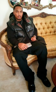 Chadric D. Henderson, dressed in black pants and a black puffy jacket, sitting on a couch holding his cell phone in hand.